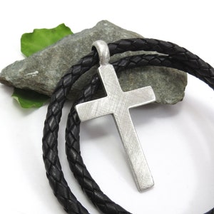 May include: A silver cross pendant on a black leather cord necklace. The cross has a textured surface and is hanging on a rock.