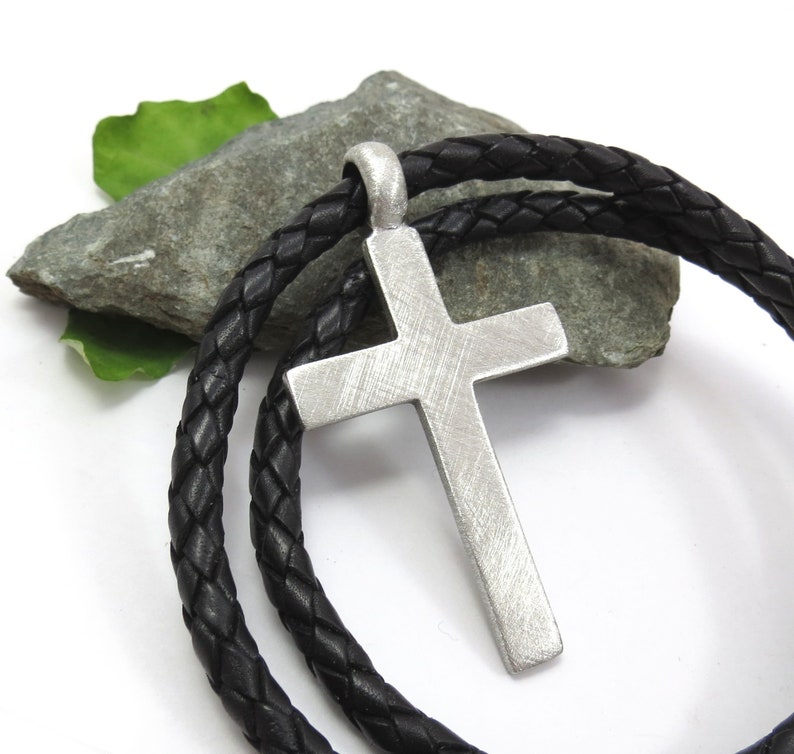 May include: A silver cross pendant on a black leather cord necklace. The cross has a textured surface and is hanging on a rock.