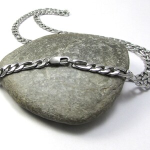 May include: A silver chain necklace with a curb link design. The chain is lying on a gray rock.