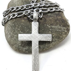 May include: A silver cross pendant with a brushed finish hangs from a silver chain. The chain and pendant are resting on a gray rock.