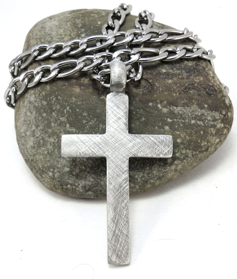 May include: A silver cross pendant with a brushed finish hangs from a silver chain. The chain and pendant are resting on a gray rock.
