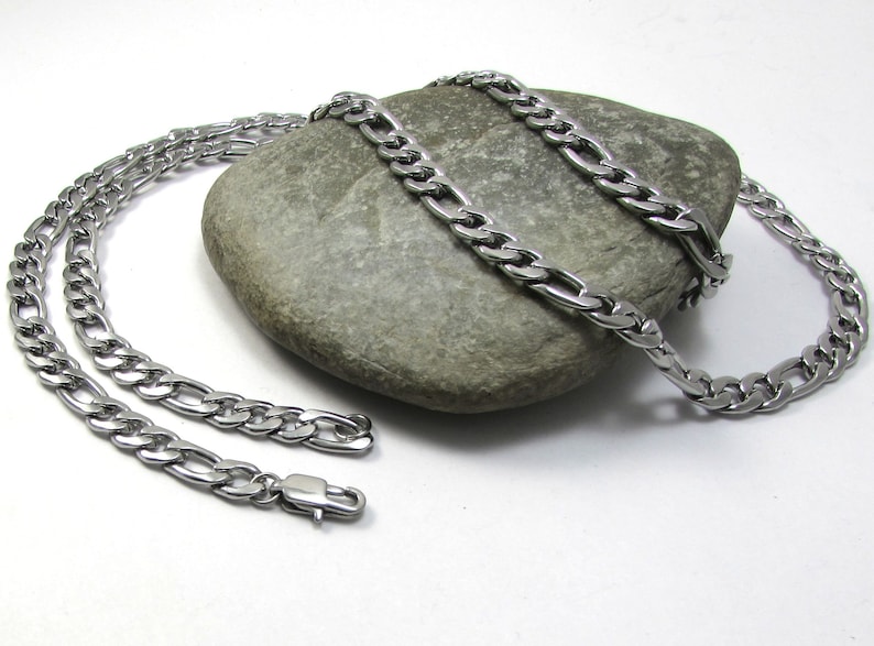 May include: Two silver curb chain necklaces with a box clasp closure. The chains are laying on a gray rock.