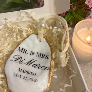 Puede incluir: Un adorno decorativo de concha de ostra con superficie blanca y bordes pintados en oro. El texto dice MR. & MRS. DiMarco MARRIED MAY 23, 2026. Incluye una cinta de terciopelo color crema con una pequeña cuenta de perla blanca, en una caja.