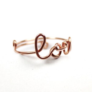 Puede incluir: Un anillo de alambre de oro rosa con la palabra "Love" escrita en cursiva.