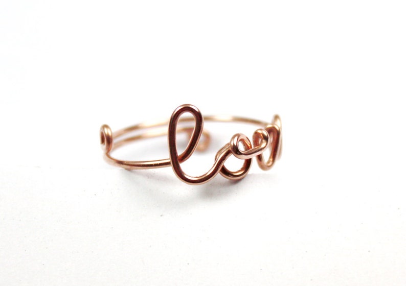 Puede incluir: Un anillo de alambre de oro rosa con la palabra "Love" escrita en cursiva.