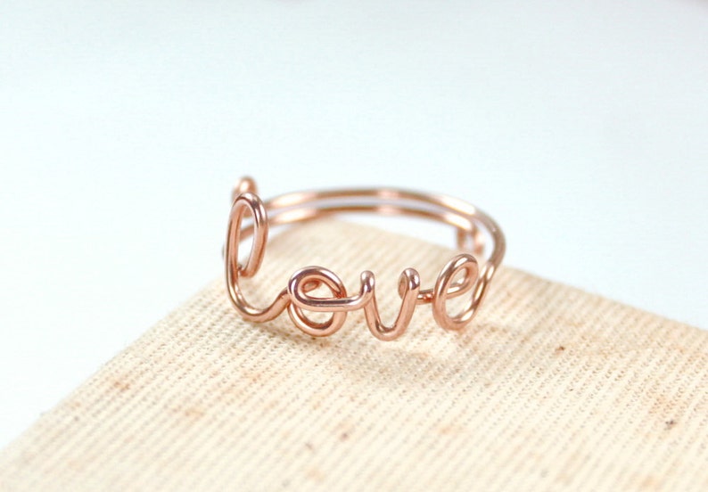 Puede incluir: Un anillo de alambre de oro rosa con la palabra "love" escrita en cursiva.