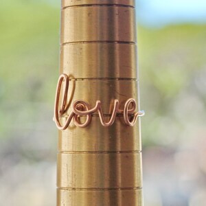 Puede incluir: Un anillo de alambre de oro rosa con la palabra "love" escrita en cursiva.