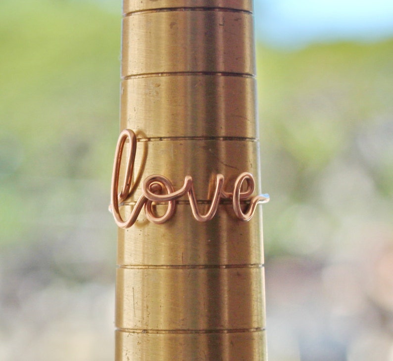 Puede incluir: Un anillo de alambre de oro rosa con la palabra "love" escrita en cursiva.