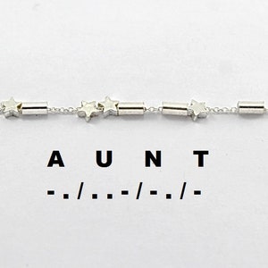 Könnte beinhalten: Ein silbernes Kettenarmband mit Stern- und Röhrenanhängern. Das Armband trägt den Schriftzug "AUNT" darunter mit Strichen und Schrägstrichen.