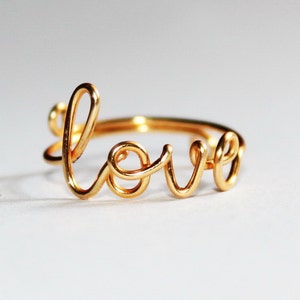 Könnte beinhalten: Ein goldfarbener Ring mit dem Wort "love" in Schreibschrift. Der Ring besteht aus einem dünnen Draht, der zu Buchstaben gebogen wurde. Der Ring liegt auf einer weißen Oberfläche.