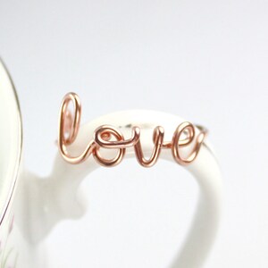 Puede incluir: Anillo de alambre de oro rosa con la palabra "love" escrita en cursiva.