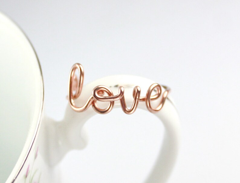 Puede incluir: Anillo de alambre de oro rosa con la palabra "love" escrita en cursiva.