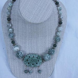 Puede incluir: Un collar de cuentas con un colgante grande tallado. El collar presenta cuentas redondas y veteadas en tonos verdes y grises, intercaladas con cuentas verdes más oscuras. El colgante tiene forma ovalada tallada con un diseño floral.