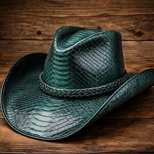 May include: A dark green cowboy hat with a snakeskin-effect texture. The hat has a braided band around the crown and a wide brim. The hat is displayed on a wooden surface.