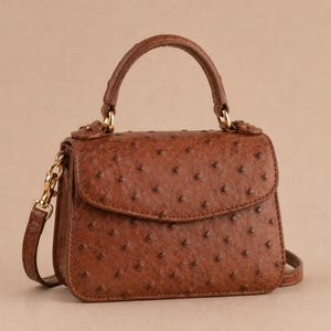 Pode incluir: Uma pequena bolsa marrom com uma superfície texturizada e uma alça superior. A bolsa possui fecho de aba e uma alça de ombro removível com ferragens douradas. A bolsa tem aproximadamente 18 cm de largura.