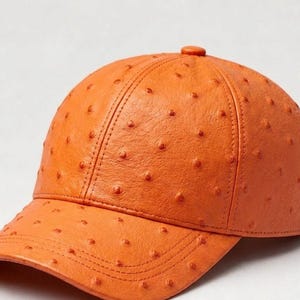 Könnte beinhalten: Orangefarbene Baseballkappe mit strukturiertem, gepunktetem Muster. Die Kappe hat einen gebogenen Schirm und einen kleinen Knopf oben. Die Nähte sind sichtbar, und die Kappe scheint aus Leder zu sein.