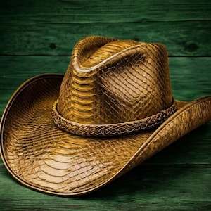 Könnte beinhalten: Ein goldbrauner Cowboyhut mit einer strukturierten, schlangenlederartigen Musterung. Der Hut hat ein geflochtenes Band um den Kopf und eine breite Krempe. Der Hut steht vor einem grünen Holzgrund.