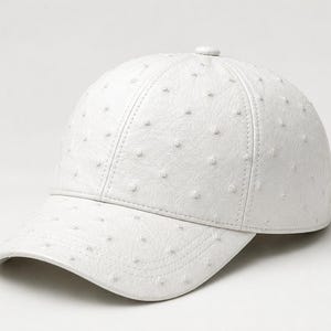 Könnte beinhalten: Eine weiße Baseballkappe mit gebogenem Schirm. Die Kappe hat eine strukturierte Oberfläche mit kleinen, gleichmäßig verteilten Erhebungen. Sie hat einen Knopf oben und besteht aus einem weichen Material.