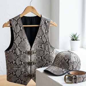 May include: A snakeskin-patterned vest, baseball cap, and belt. The vest has a V-neck and three buckle closures. The cap has a curved brim. The belt has a brass buckle. The items are displayed on a white surface.