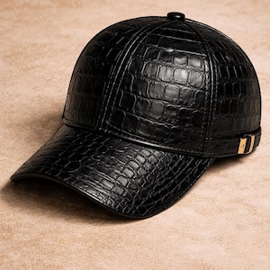 Könnte beinhalten: Schwarze Baseballkappe mit strukturiertem, krokodilähnlichem Muster. Die Kappe hat einen gebogenen Schirm und einen kleinen, runden Knopf oben. Ein goldfarbener Akzent ist an der Seite sichtbar.