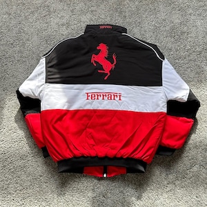 May include: A black, white, and red Ferrari racing jacket. The jacket features a black upper back with a red Ferrari logo and a white stripe across the chest. The Ferrari logo is also displayed across the chest in red.