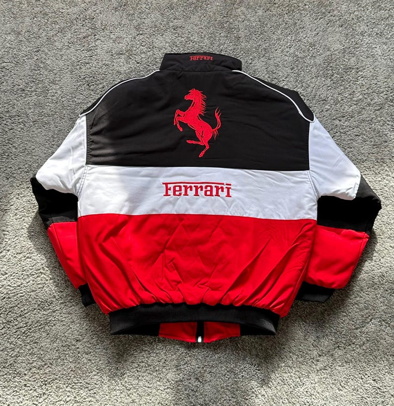 May include: A black, white, and red Ferrari racing jacket. The jacket features a black upper back with a red Ferrari logo and a white stripe across the chest. The Ferrari logo is also displayed across the chest in red.