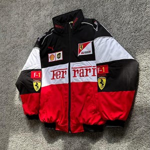May include: Black, white, and red Ferrari racing jacket with multiple logos, including Puma and Shell. The jacket features a full-zip closure, a stand-up collar, and the word "Ferrari" prominently displayed across the chest.