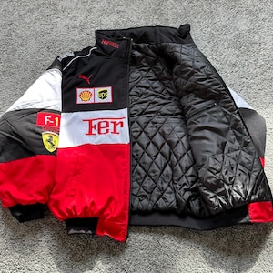 May include: A black, red, and white Ferrari racing jacket with the Ferrari logo, Shell, and UPS logos. The jacket has a quilted black interior and the word "Fer" in large white letters on the front.