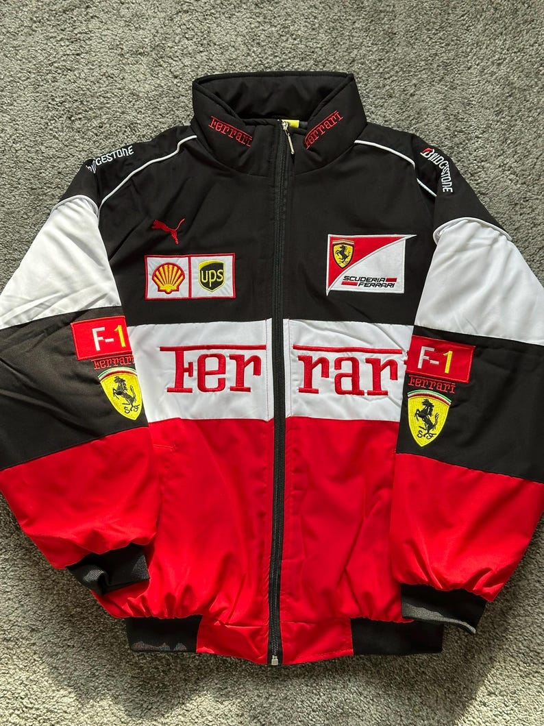 May include: Black, red, and white Ferrari racing jacket with the Ferrari logo and text. The jacket features a zip-up front, a stand-up collar, and various sponsor logos. The sleeves have black cuffs and white panels.