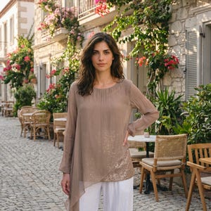 Puede incluir: Una mujer con una blusa marrón claro de manga larga con un dobladillo asimétrico con lentejuelas y pantalones blancos. La blusa tiene una tela transparente y fluida. El fondo muestra una calle adoquinada, asientos al aire libre y edificios con enredaderas florecientes.