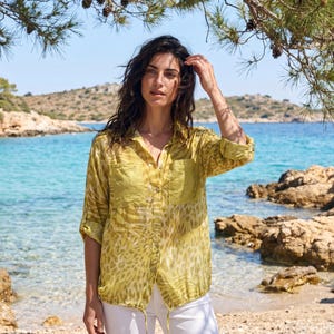 Puede incluir: Una camisa amarilla con botones y un diseño de estampado animal único. La camisa tiene dos bolsillos delanteros y mangas enrolladas. La persona lleva pantalones blancos. El fondo presenta una playa con agua turquesa y cielo azul claro.