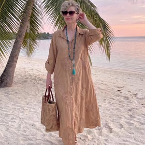 Può includere: Abito camicia beige con ricami floreali, borsa tote intrecciata e lunga collana di perline turchesi. L'abito è al ginocchio e ha maniche arrotolate. La donna indossa occhiali da sole e sandali su una spiaggia sabbiosa.