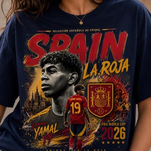 以下が含まれることがあります： サッカー選手ラミン・ヤマルがプリントされたネイビーブルーのTシャツ。デザインには「SPAIN」「LA ROJA」「Lamine YAMAL 19」「FIFA WORLD CUP 2026」の文字、スペイン代表チームのエンブレム、赤いユニフォームを着た選手の姿が描かれています。