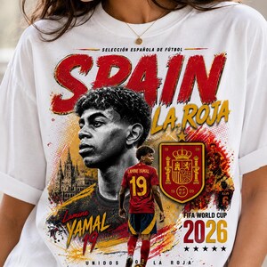 Può includere: Una t-shirt bianca con una stampa grafica di Lamine Yamal, lo stemma della nazionale di calcio spagnola e il testo "SPAIN," "LA ROJA," "Lamine YAMAL 19," "SELECCIÓN ESPAÑOLA DE FÚTBOL," "FIFA WORLD CUP 2026," e "UNIDOS POR LA ROJA."