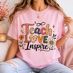 Pode incluir: Uma t-shirt de manga curta rosa claro com o texto Teach Love Inspire numa fonte estampada multicolorida. O design inclui uma forma de coração, várias ilustrações florais e pequenos detalhes de estrelas em tons de vermelho, laranja, verde e amarelo.