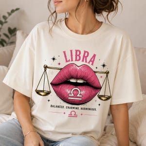 May include: A cream-colored t-shirt featuring a graphic of pink lips centered within a gold scale of justice. The text LIBRA appears above the lips, while the words BALANCED, CHARMING, HARMONIOUS and the Libra zodiac symbol are printed below.