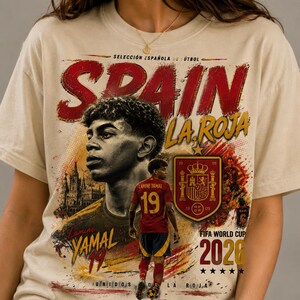 以下が含まれることがあります： サッカー選手ラミン・ヤマル、テキスト「SPAIN LA ROJA」、「Lamine YAMAL 19」、「FIFA WORLD CUP 2026」、「UNIDOS POR LA ROJA」のグラフィックプリントが施されたベージュのTシャツ。デザインにはスペイン代表チームのエンブレムと大聖堂が含まれています。