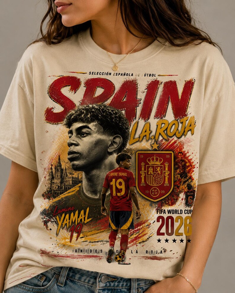 以下が含まれることがあります： サッカー選手ラミン・ヤマル、テキスト「SPAIN LA ROJA」、「Lamine YAMAL 19」、「FIFA WORLD CUP 2026」、「UNIDOS POR LA ROJA」のグラフィックプリントが施されたベージュのTシャツ。デザインにはスペイン代表チームのエンブレムと大聖堂が含まれています。