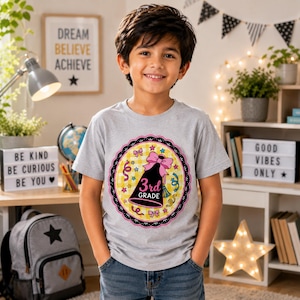 Può includere: Una t-shirt grigio chiaro a maniche corte con una grafica circolare su sfondo a quadretti gialli, un bordo smerlato nero e un'illustrazione centrale di un megafono nero con un fiocco rosa e la scritta 3rd GRADE stampata in bianco.