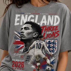 Puede incluir: Una camiseta gris con un estampado gr&aacute;fico de Jude Bellingham. El dise&ntilde;o incluye el texto "ENGLAND", "THREE LIONS", "Jude Bellingham 10", "FIFA WORLD CUP 2026" y "GOD SAVE THE KING", junto a un jugador de f&uacute;tbol, un escudo y un bal&oacute;n.