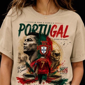May include: A beige t-shirt featuring a graphic print of a soccer player, the Portugal national team crest, and the FIFA World Cup 2026 trophy. The design includes the text PORTUGAL, RUMO À GLÓRIA, SELEÇÃO DAS QUINAS, Por Amor À Camisola, and 2026.