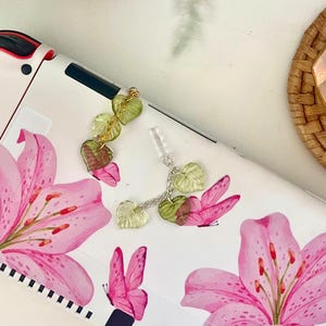 May include: White case with pink lily and butterfly graphics. A silver chain with green leaf charms is attached. A potted plant and a candle holder are in the background.