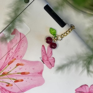 Op de afbeelding: Witte elektronische apparaathoes met een bloemen- en vlinderontwerp. De hoes heeft een roze lelie en roze vlinders. Een gouden ketting met rode kersenbedels hangt aan het apparaat. De hoes is versierd met een zwart-witte gestreepte rand.