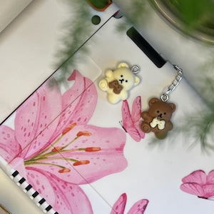 Könnte beinhalten: Eine weiße Hülle mit rosa Blumenmuster und zwei Teddybär-Anhängern. Ein Anhänger ist cremefarben mit einem braunen Bären, der andere ist braun. Die Hülle zeigt rosa Lilien und Schmetterlinge.