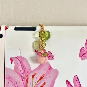 Op de afbeelding: Een goudkleurige bedel met een roze appel en groene bladeren. De bedel is bevestigd aan een gouden ketting. De achtergrond is een wit oppervlak met roze bloemen- en vlindermotieven.
