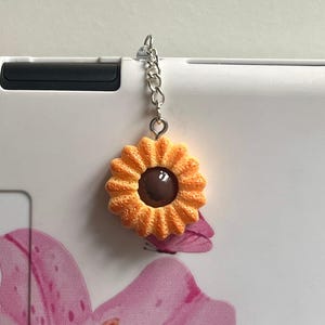 May include: A sunflower-shaped charm with a dark brown centre and orange petals, attached to a silver chain. The charm is hanging from a white surface, with a pink floral design in the background.
