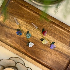 Peut inclure: Deux boucles d'oreilles fleuries avec des chaînes dorées et des breloques en forme de fleurs colorées. Les boucles d'oreilles présentent des fleurs bleues, vertes, blanches, jaunes, rouges et violettes avec de petits accents de feuilles vertes. Elles sont présentées sur un plateau en bois.