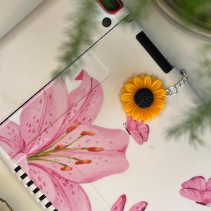 Puede incluir: Funda blanca para dispositivo electrónico con diseño floral y mariposas rosas. Un charm de girasol amarillo con centro negro está sujeto con una cadena plateada. La funda tiene un borde a rayas blanco y negro.