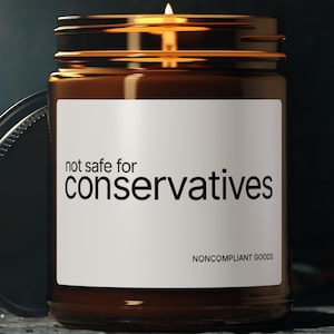 Może przedstawiać: Zapalone świeca w brązowym szklanym słoiku ze złotą pokrywką. Etykieta głosi "not safe for conservatives" i "NONCOMPLIANT GOODS". Słoik stoi na ciemnej powierzchni.