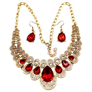 May include: A gold-toned necklace and earring set. The necklace features large, teardrop-shaped red gemstones surrounded by clear crystals. The earrings mirror the teardrop design. The chain is a linked style.
